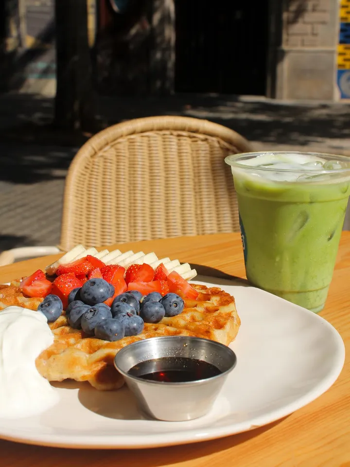 Waffle con fresas, arándanos y yogur junto a un iced matcha latte, en una mesa al sol