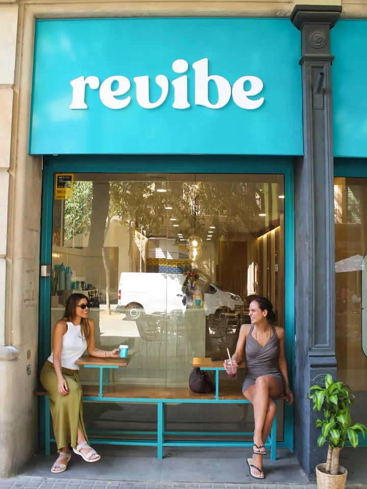 La fachada turquesa de Revibe en Carrer de Bailèn, con dos clientas tomando algo en el banco de la entrada